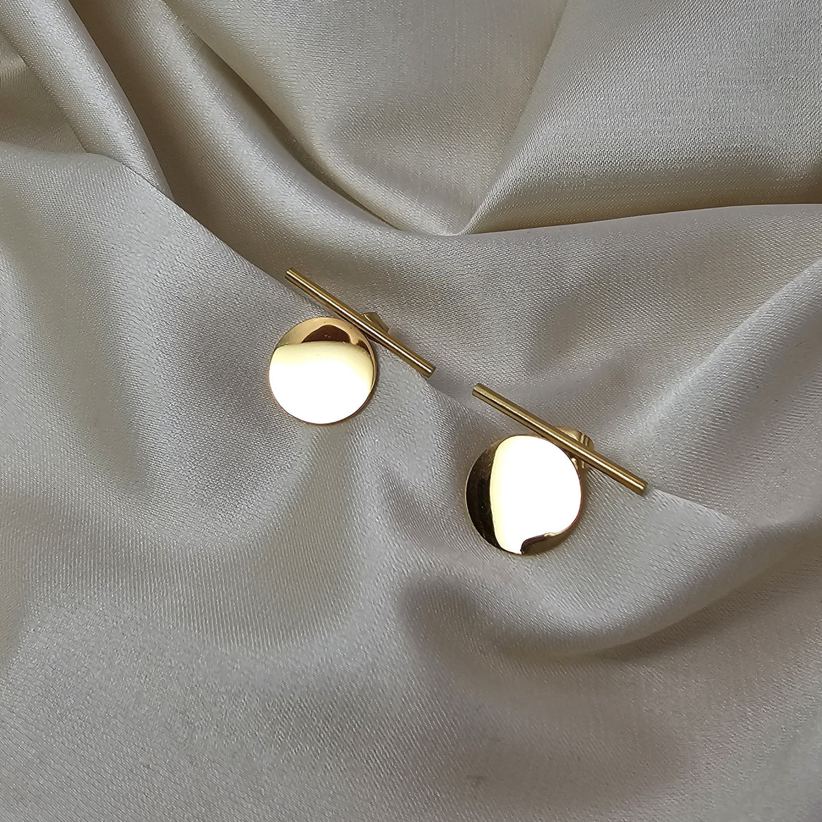 A pair of gold-toned stud earrings with circular disc fronts and straight post backs, displayed on draped cream satin fabric.