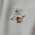 A silver spider pendant featuring a central amber-colored gemstone, displayed on a textured light gray fabric background.