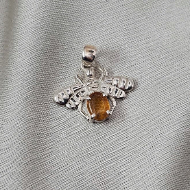 A silver spider pendant featuring a central amber-colored gemstone, displayed on a textured light gray fabric background.