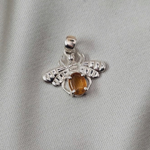 A silver spider pendant featuring a central amber-colored gemstone, displayed on a textured light gray fabric background.