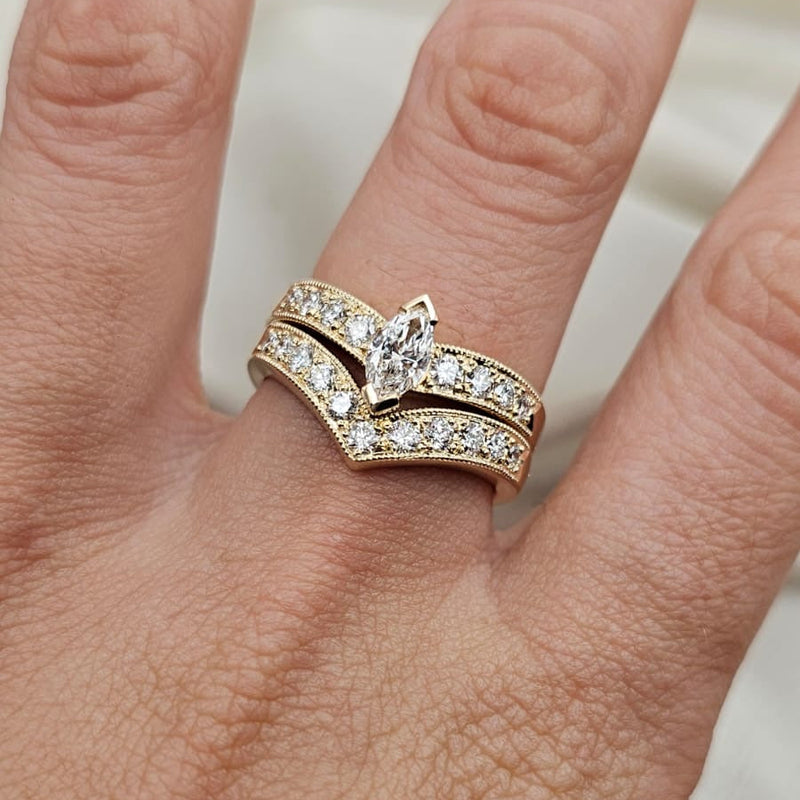 A close-up of a hand wearing a two-piece yellow gold wedding ring set. The set features a V-shaped band with pavé-set round diamonds and a matching marquise-cut diamond center stone.